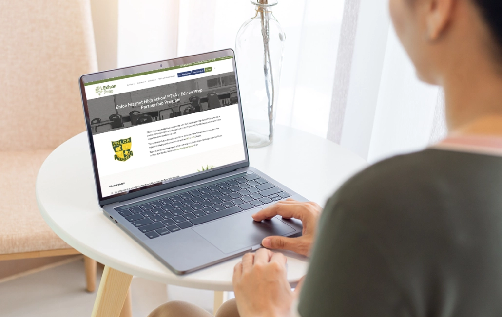 Close-up of a person using a laptop to view a webpage for the Enloe Magnet High School PTSA/Edison Prep Partnership Program. The screen shows details about the program, including the Enloe Eagles crest and information about included services. The laptop rests on a white table with a glass vase in the background.